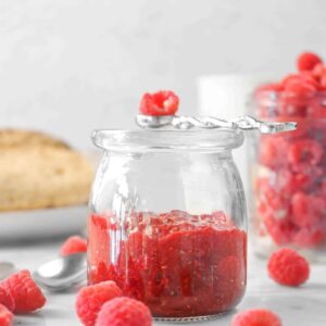 raspberry jam in a glass jar with fresh raspberries, bread, and a jar of raspberries in the background