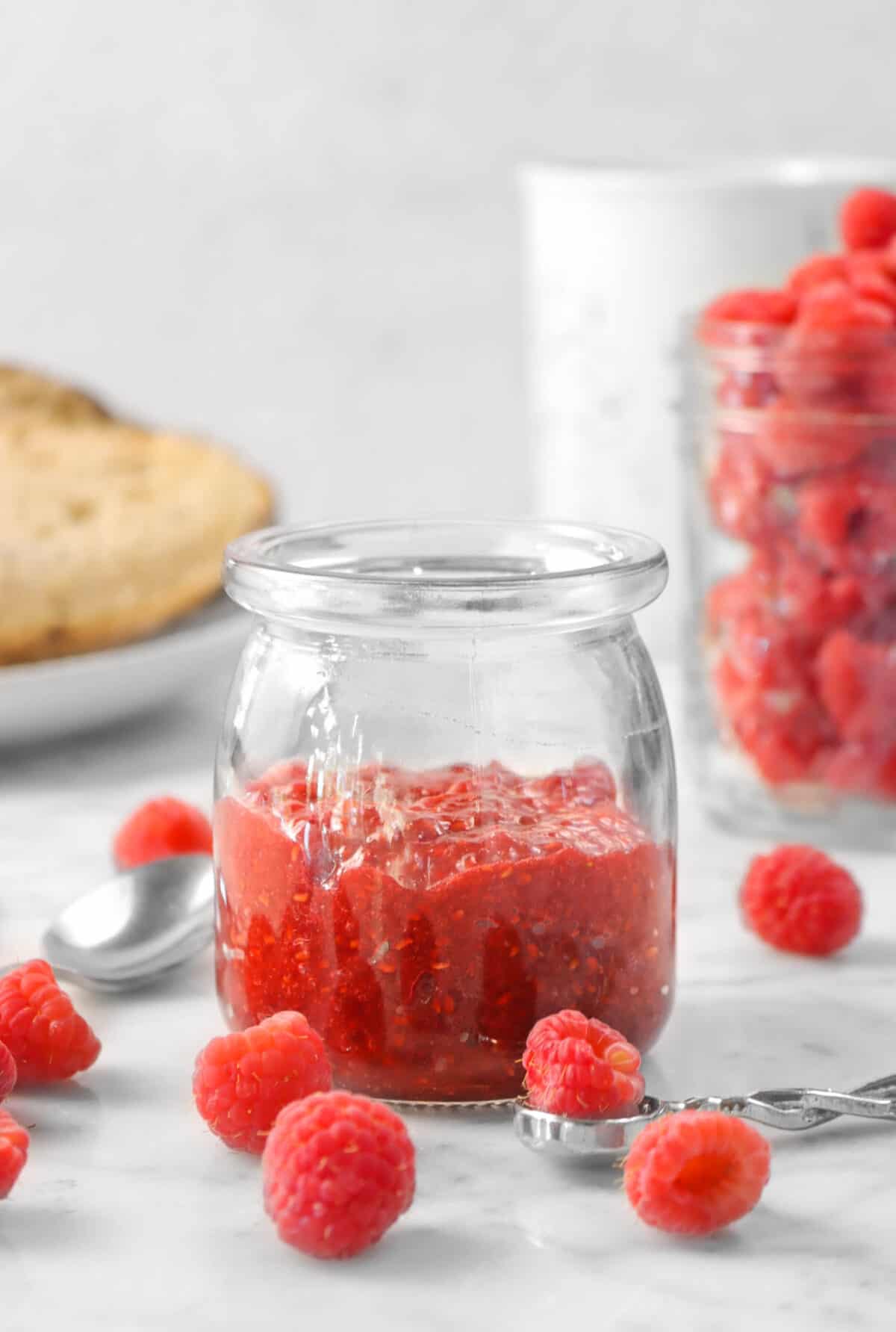 a jar of raspberry jam with two spoons, fresh raspberries, and toast