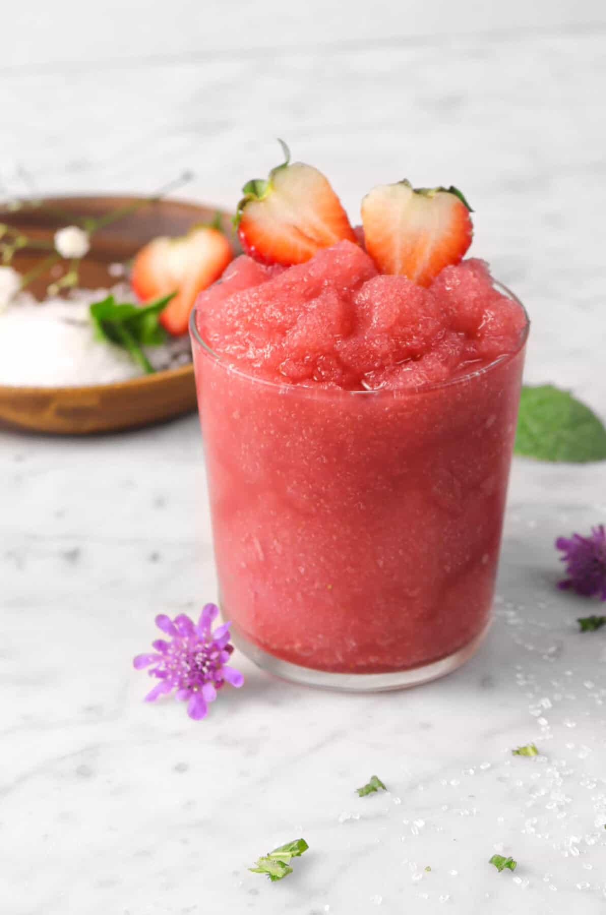 strawberry moscato slush in a glass with fresh strawberries, flowers, mint, sugar, and a wood plate
