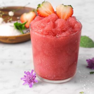 strawberry moscato slush in a glass with fresh strawberries, flowers, mint, sugar, and a wood plate