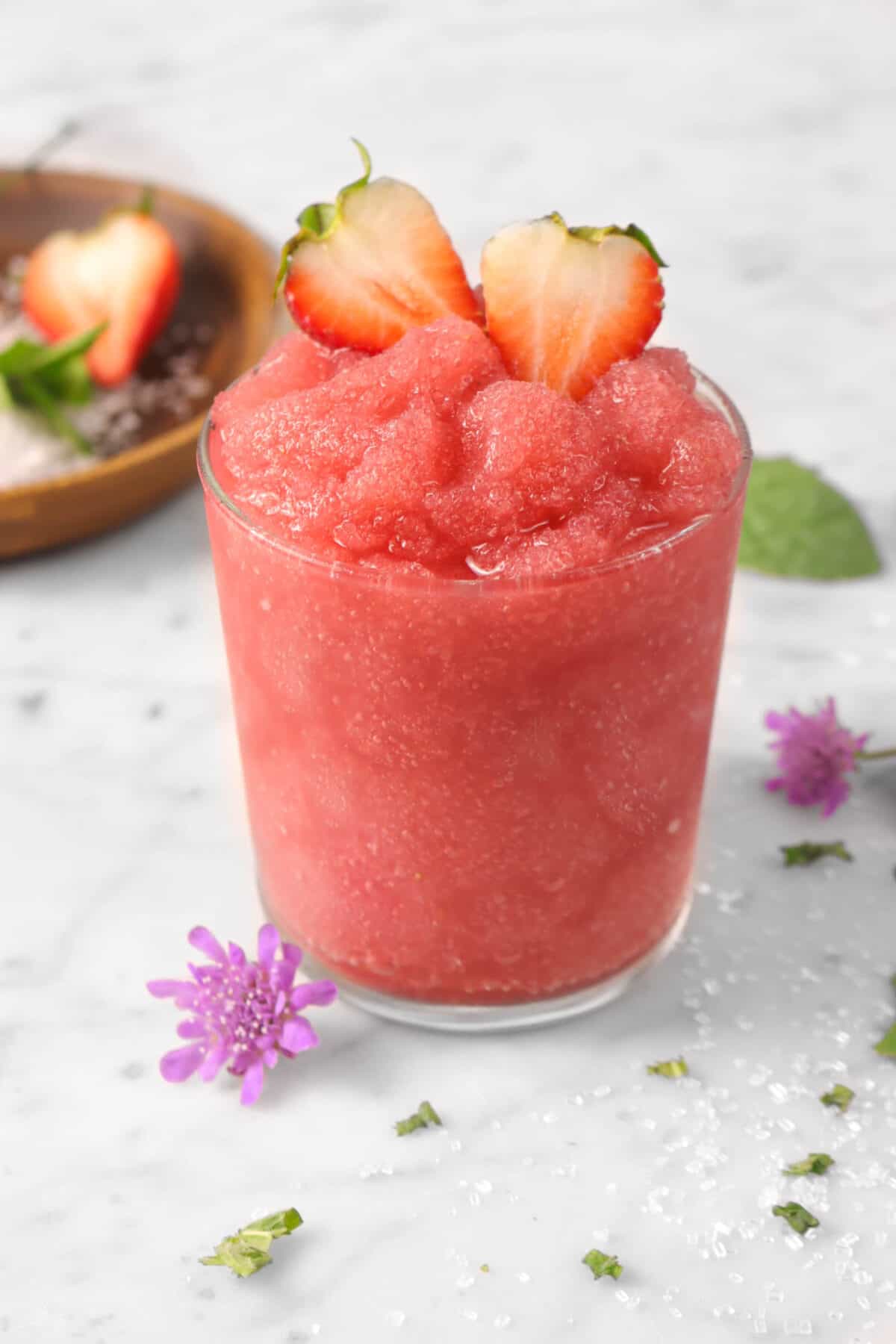 a glass of strawberry slush with mint, strawberries, purple flowers, and sugar