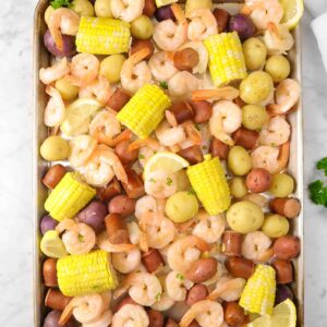 shrimp boil on a sheet pan with a glass of beer, a can, a small bowl of spices, and a white napkin