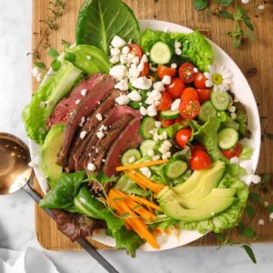 steak salad with white flowers, herbs, a copper serving spoon and a white napkin on a wood board