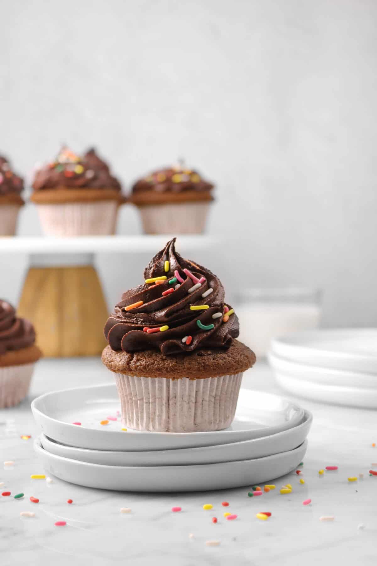 a cupcake on three plates with more cupcakes behind and sprinkles