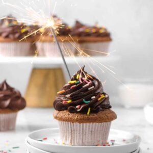chocolate cupcake on white plates with sprinkles, a sparkler, and more cupcakes behind