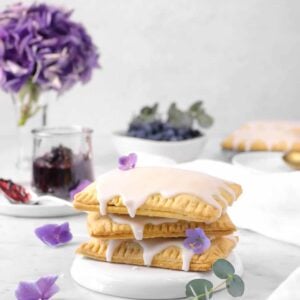 three pop tarts stacked with purple flowers, a napkin, a jar of jam, eucalyptus, a bowl of blueberries, and more pop tarts behind