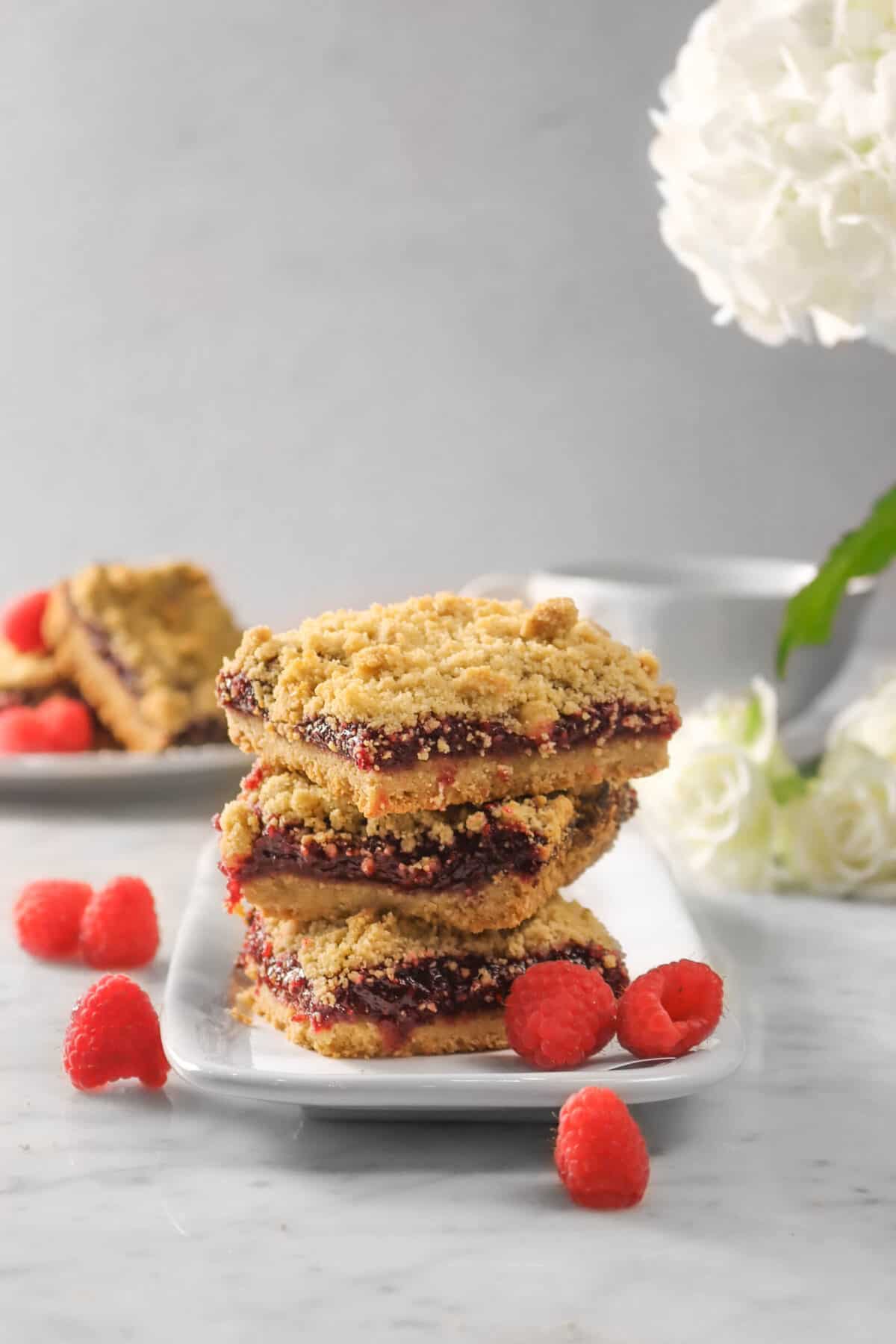 three raspberry crumble bars stacked on white plate with raspberries, fresh flowers, and more bars behind