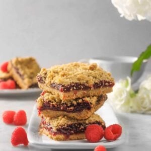three raspberry crumble bars stacked on white palte with raspberries, fresh flowers, and more bars behind