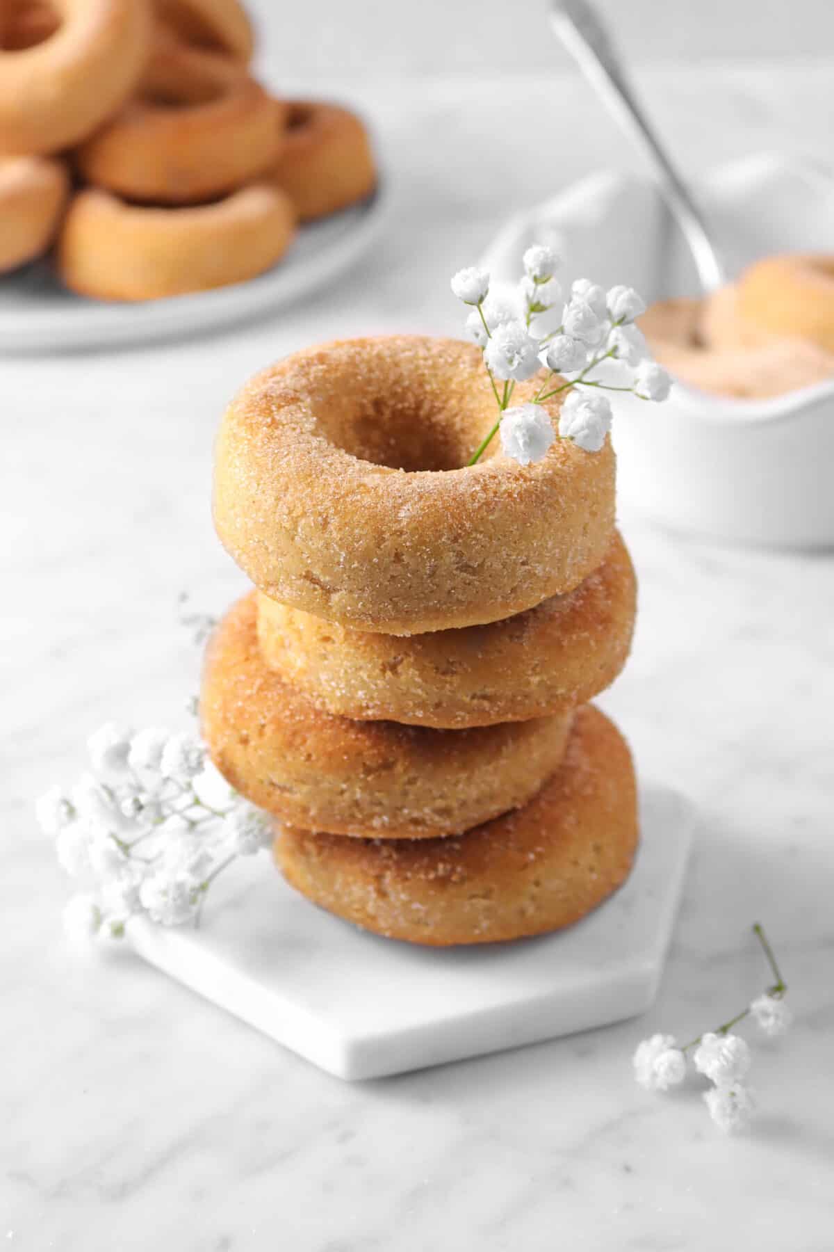 four cinnamon donuts stacked on a marble coaster with flowers, cinnamon sugar, and more donuts behind