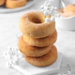 four cinnamon donuts stacked on a marble coaster with flowers, cinnamon sugar, and more donuts behind