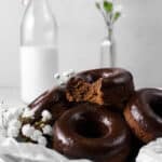 chocolate donuts with a bite out of one in a silver baking pan, parchment paper, jug of milk, and flowers