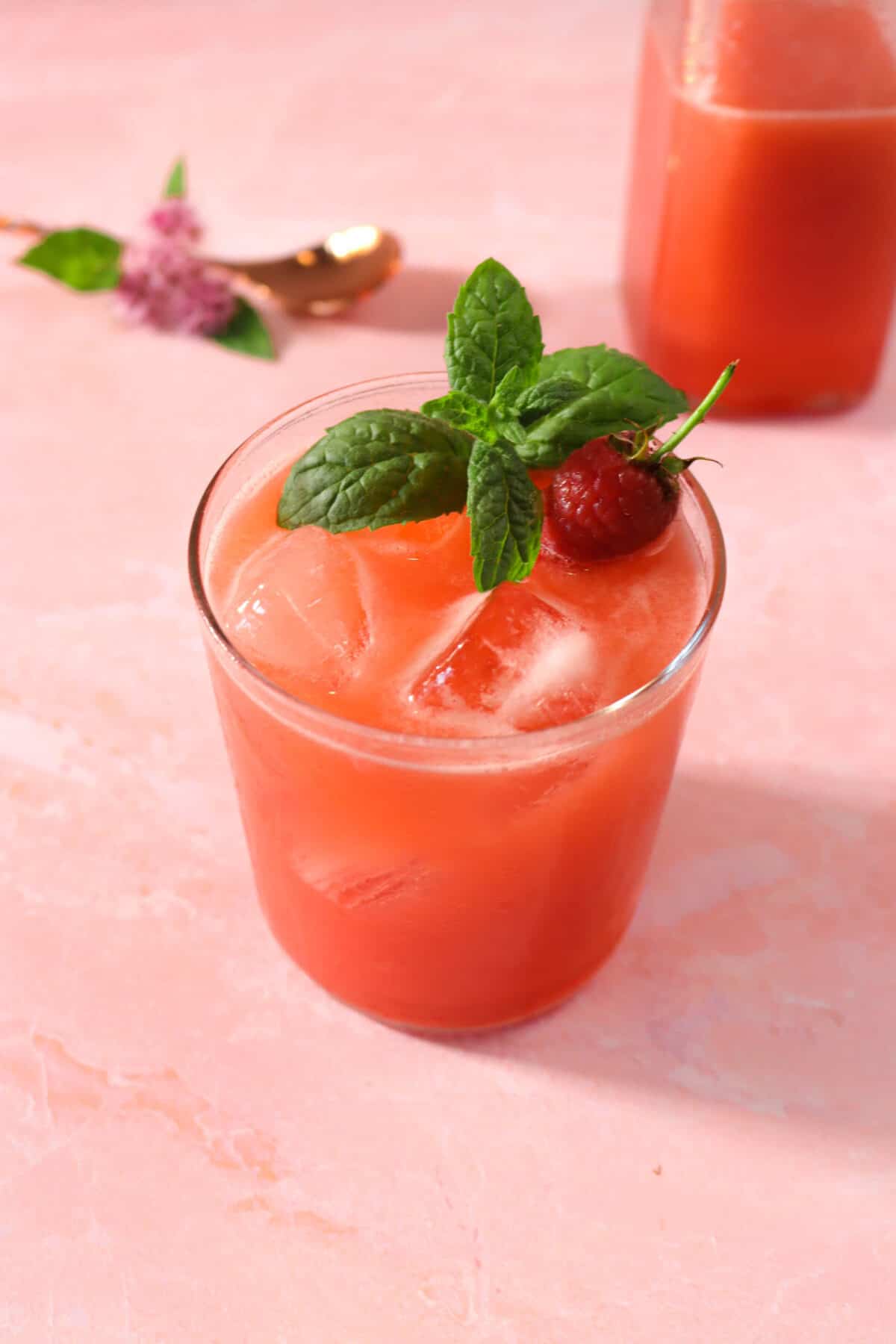 a glass of raspberry lemonade with a mint sprig, pink flowers, and a copper spoon