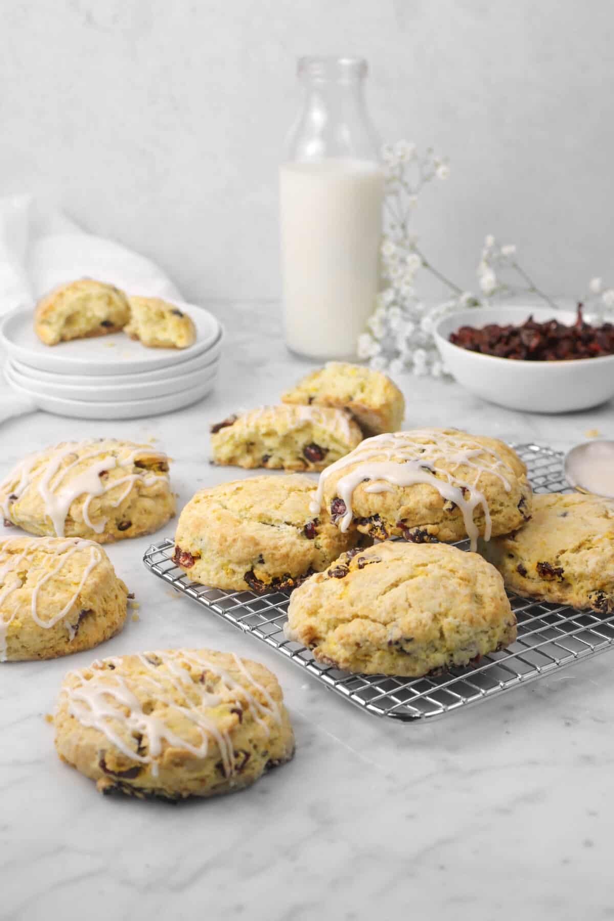 four scones on a cooling rack with three beside, a glass of milk, bowl of cranberries, and flowers behind