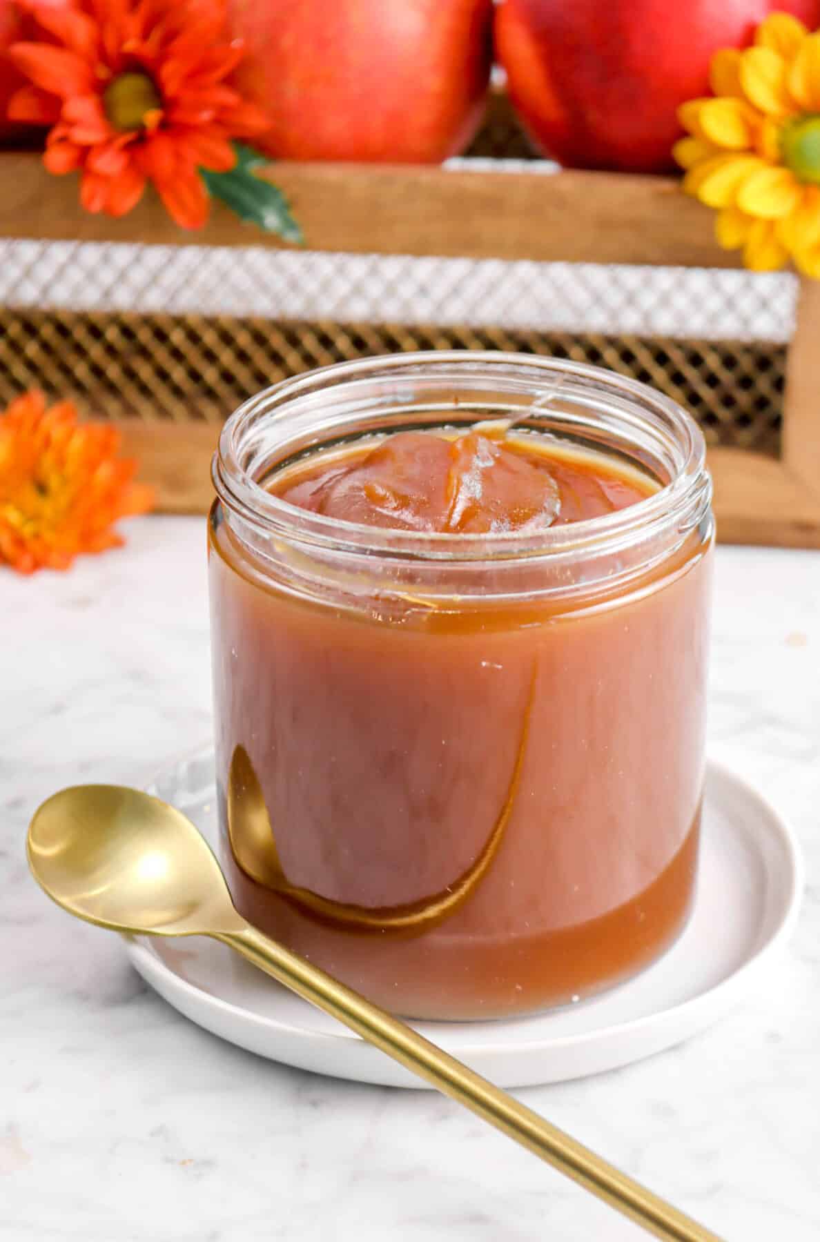 apple butter in a glass jar on a white plate with a gold spoon, flowers, and apples behind