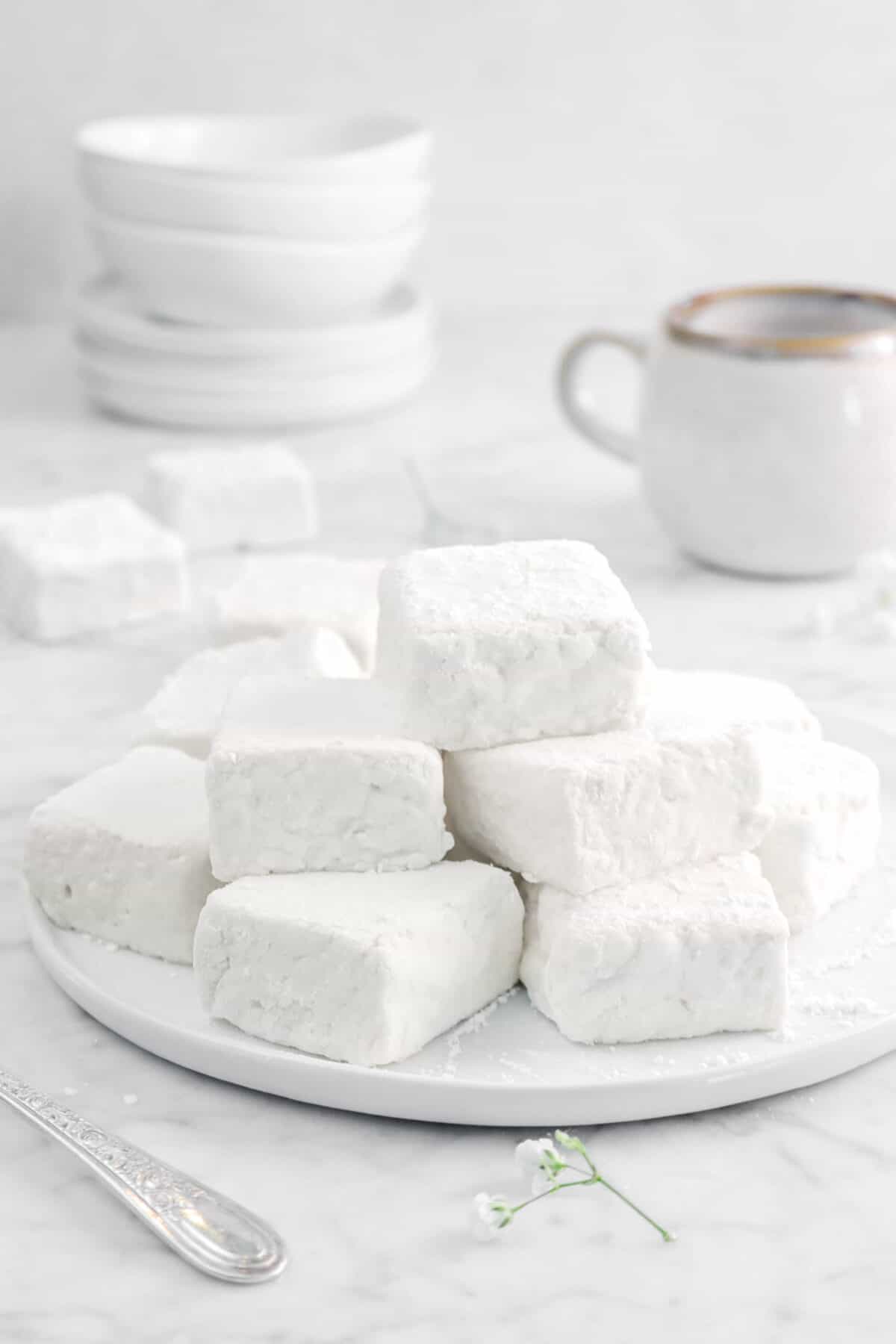 marshmallows stacked on a white plate with a mug and plates behind