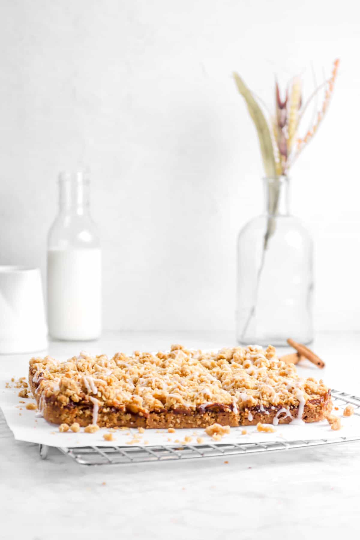 crumble bars on a cooling rack with milk, wheat, and cinnamon sticks behind