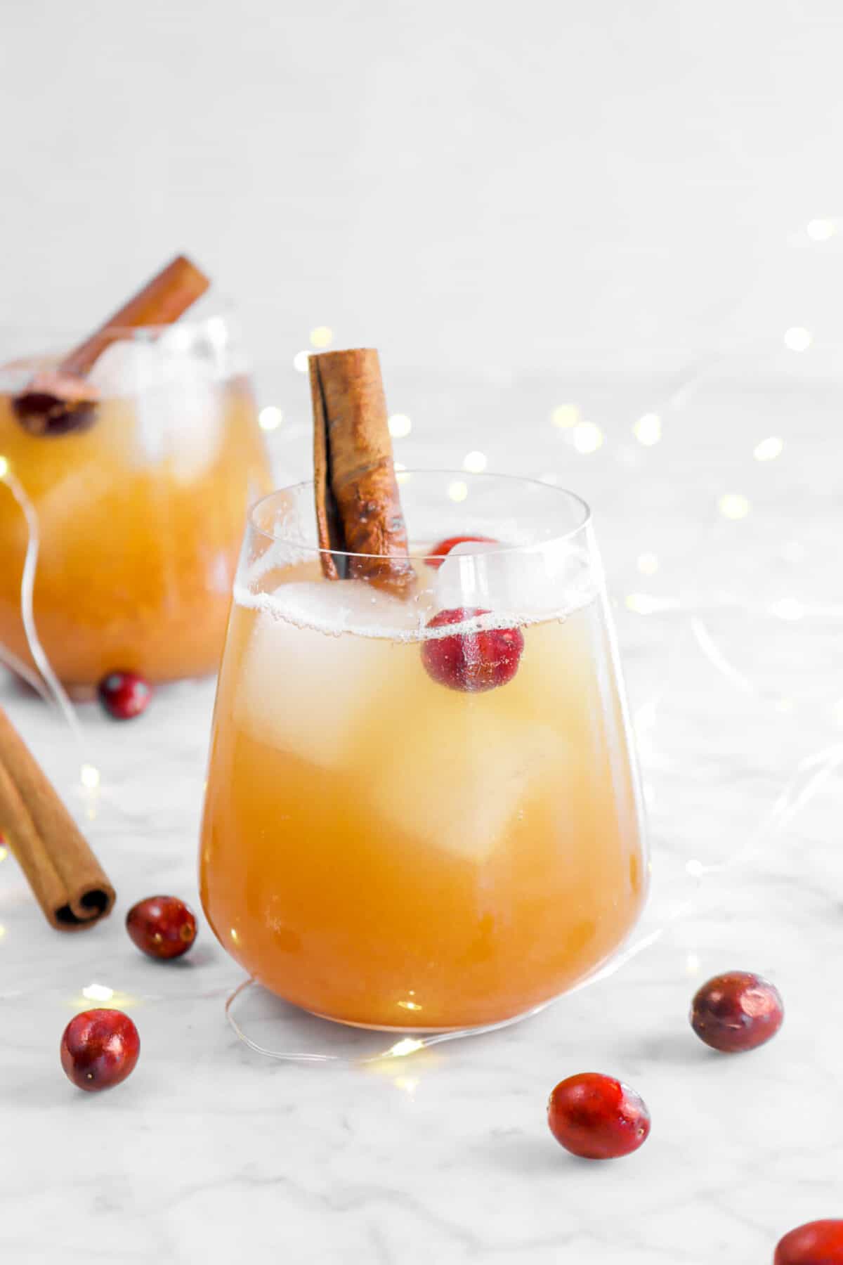 two glasses of apple cocktail with cranberries and fairy lights behind