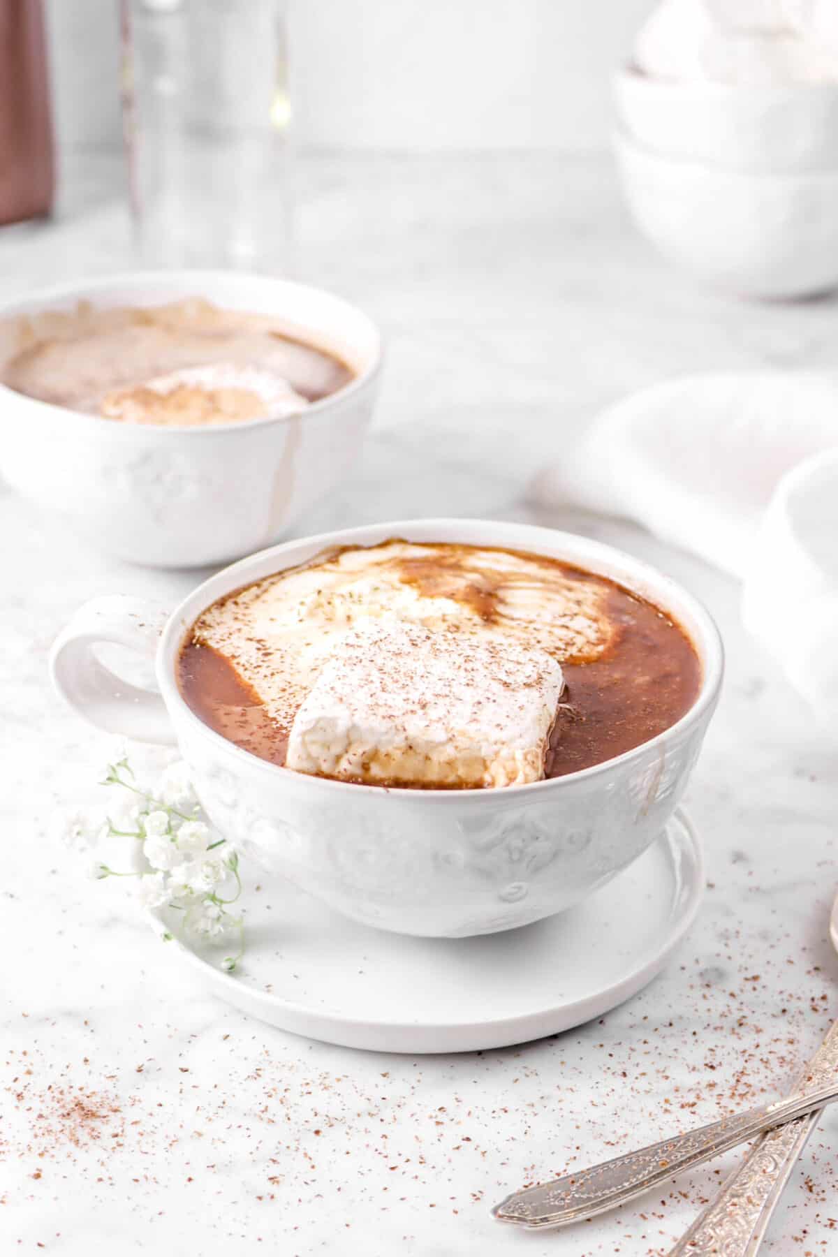 two mugs of hot chocolate with grated chocolate, marshmallows, and two spoons
