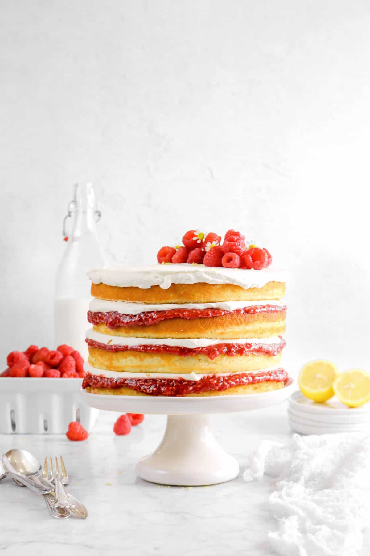 lemon raspberry jam cake on white cake plate with cheese cloth, forks, sliced lemon, raspberries, and milk