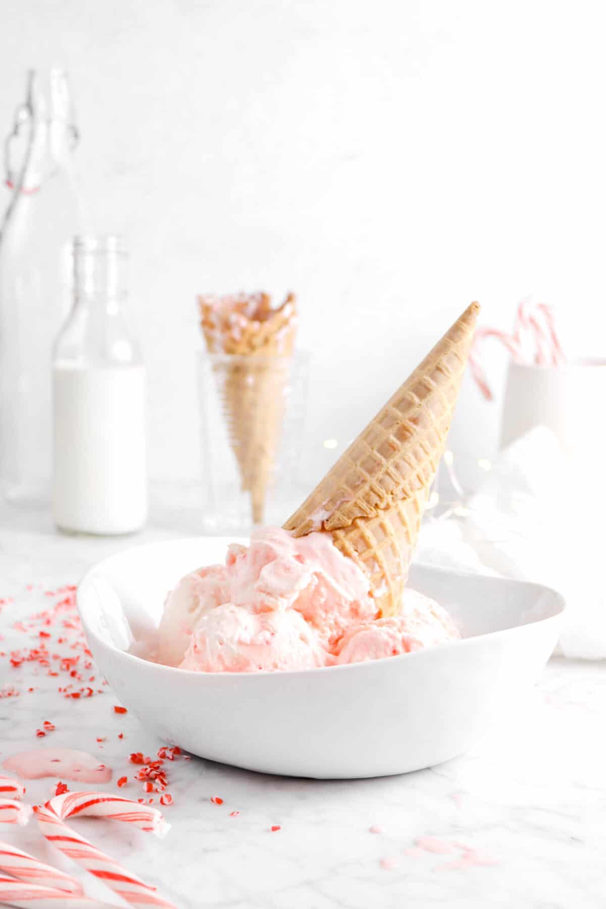 peppermint ice cream in white bowl with a cone, crushed peppermint, glass of milk, and more cones behind