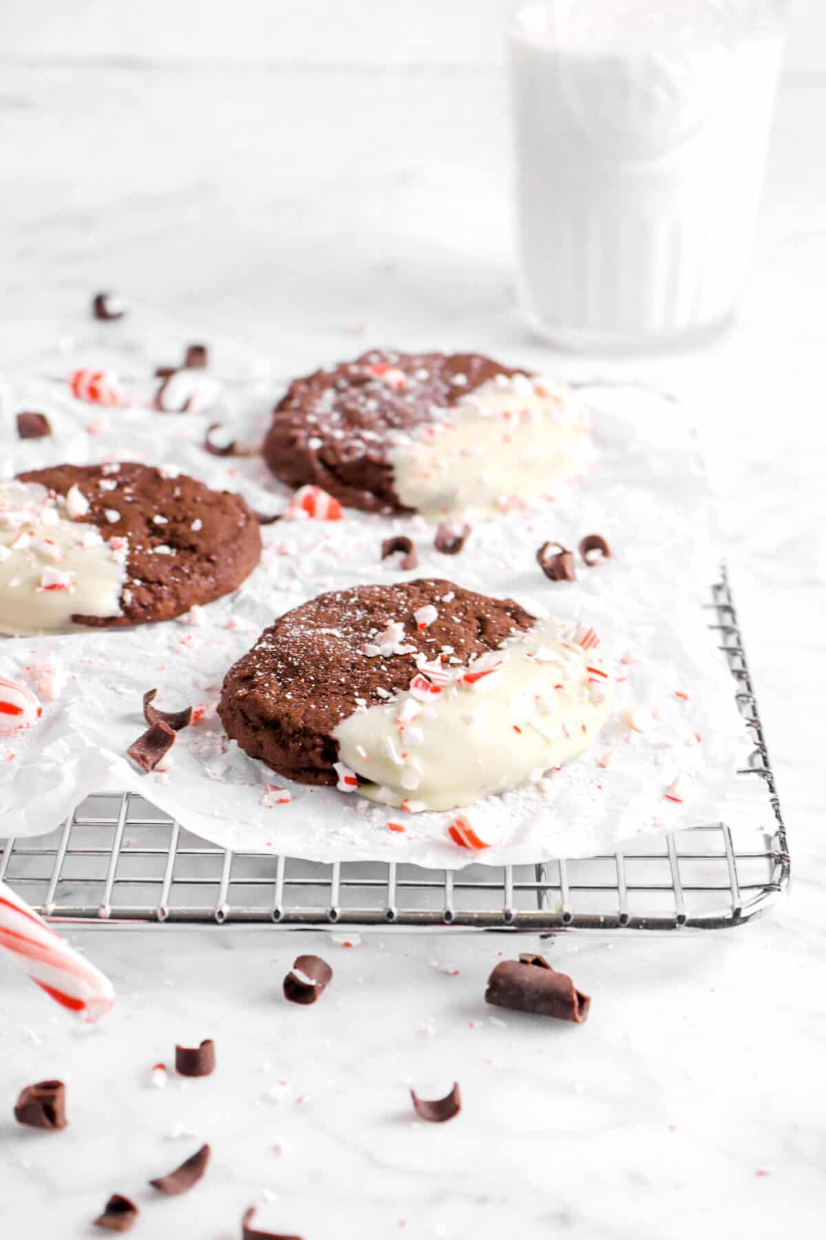 three chocolate peppermint cookies on wire cooling rack with glass of milk behind, chocolate curls, and crushed peppmint