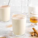 eggnog in two glasses with nutmeg on top with milk, bourbon, and cinnamon sticks