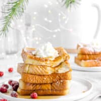 french toast on a white plate with greenery, more toast behind, fairy lights, and cranberries