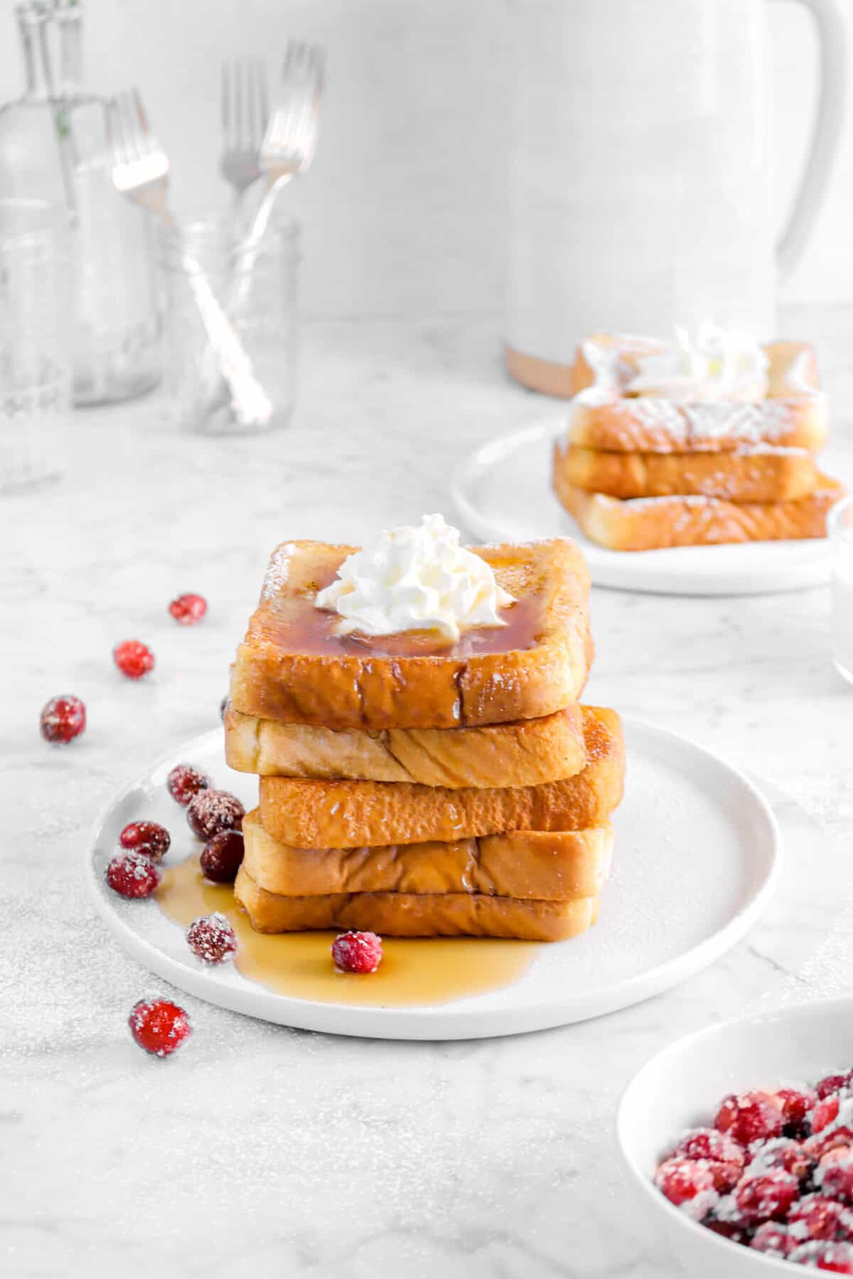 french toast on a white plate with more behind, cranberries, forks, and a pitcher