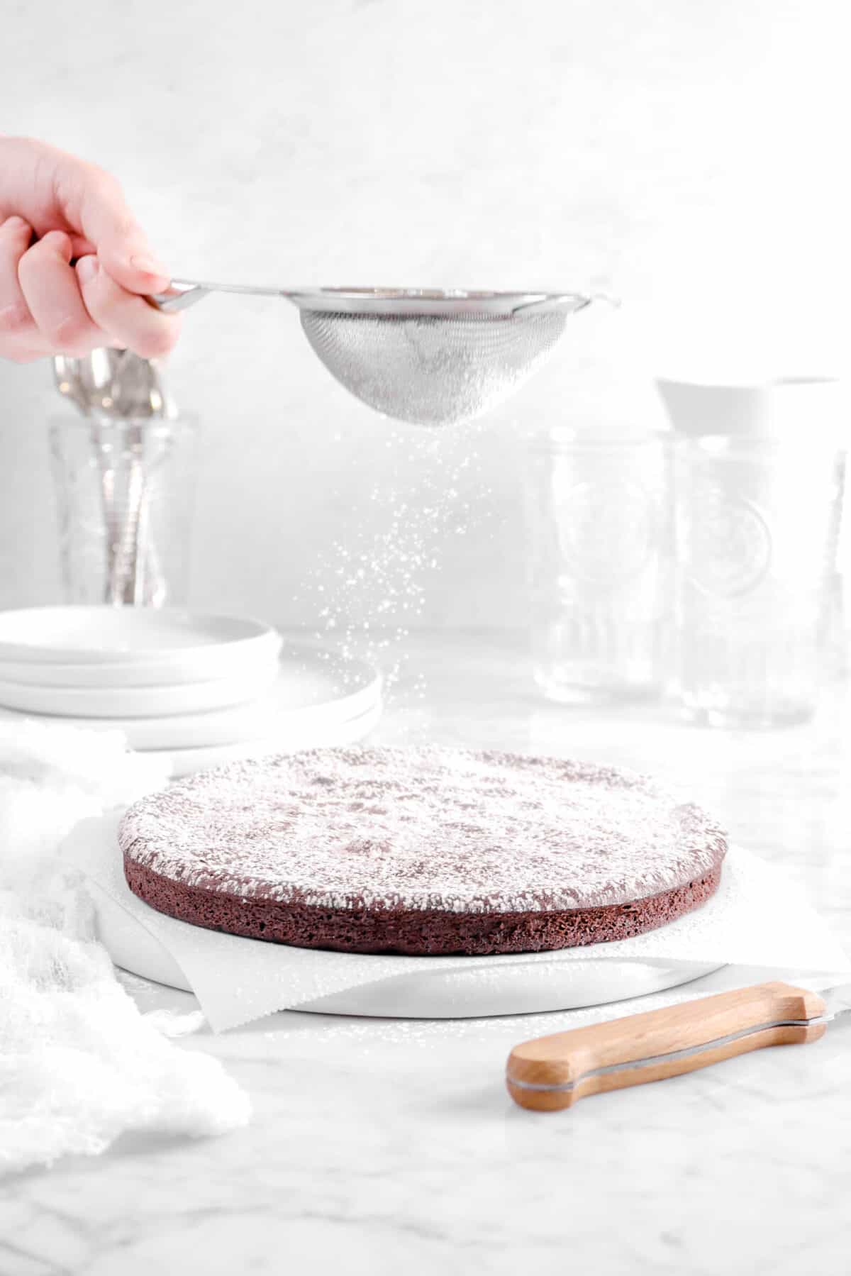 powdered sugar being sifted onto cake with glasses, plates, utensils, and a knife