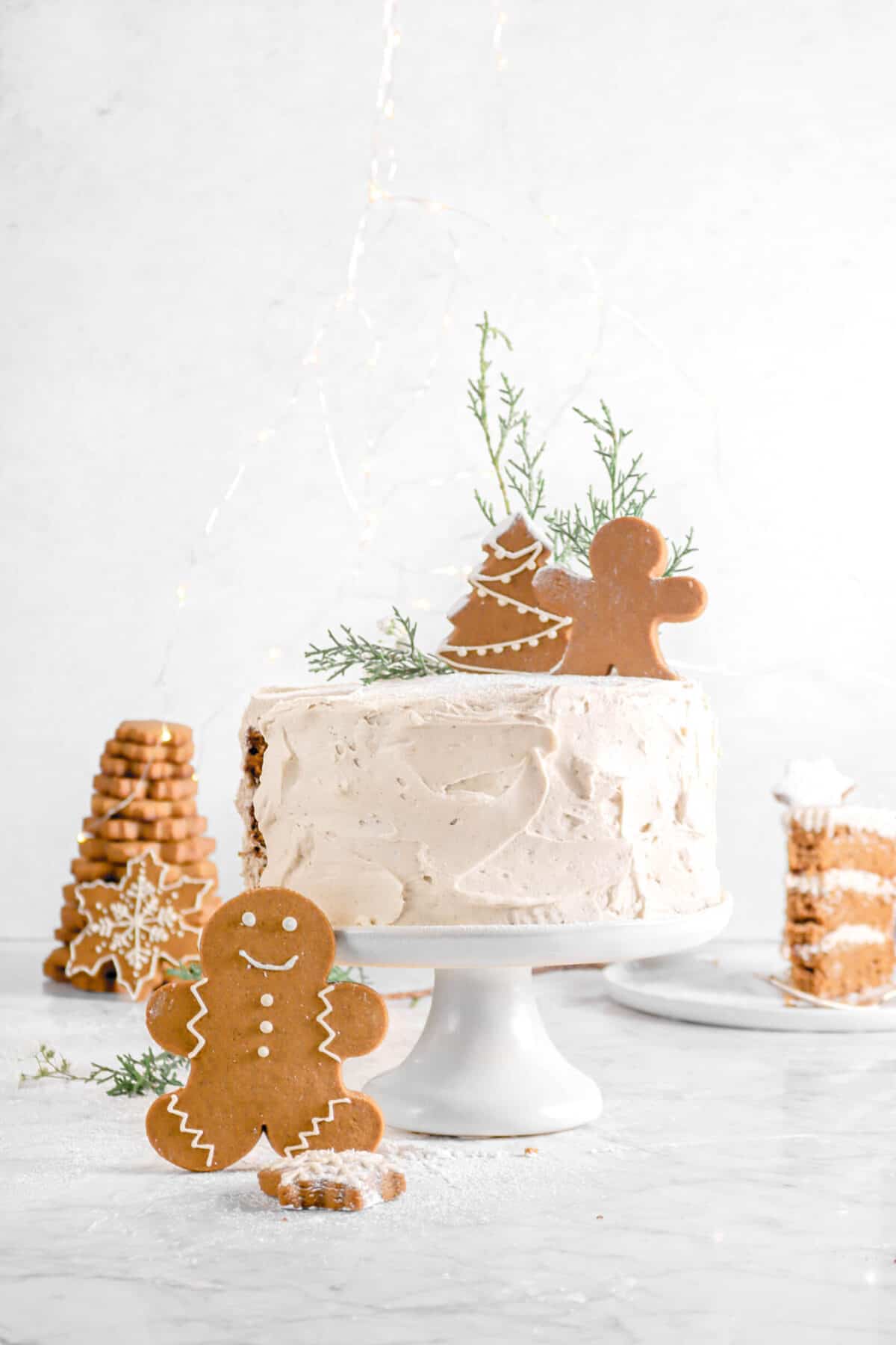 slice taken out of gingerbread cake with slice behind, cookies, greenery, and fairy lights