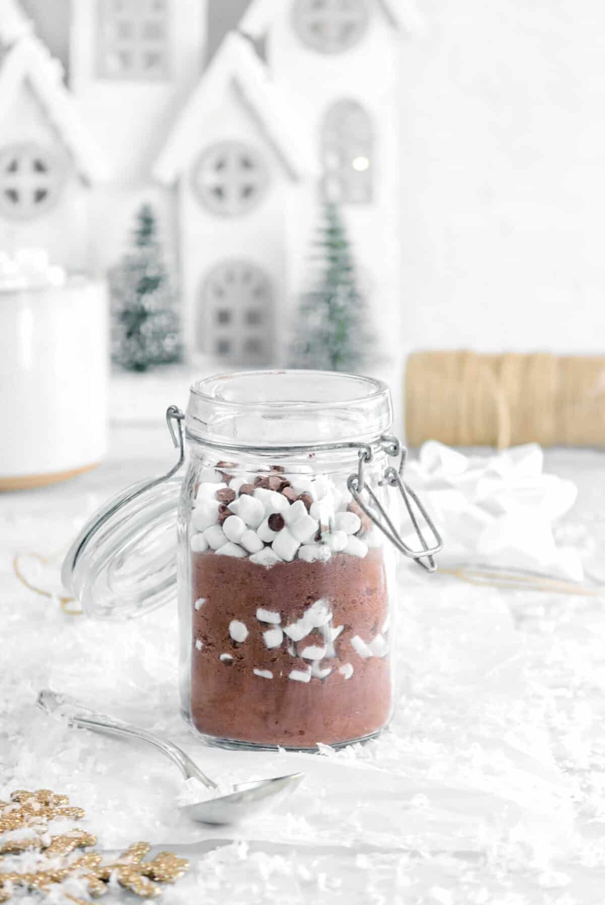 hot chocolte mix in a jar with mug, white house, green trees, and twine behind