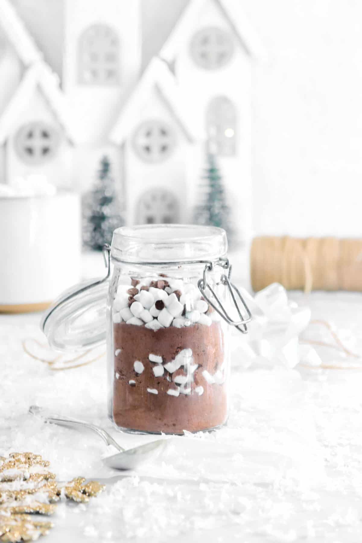 glass jar of cocoa mix with a gold slow flake, a mug, and white houses behind