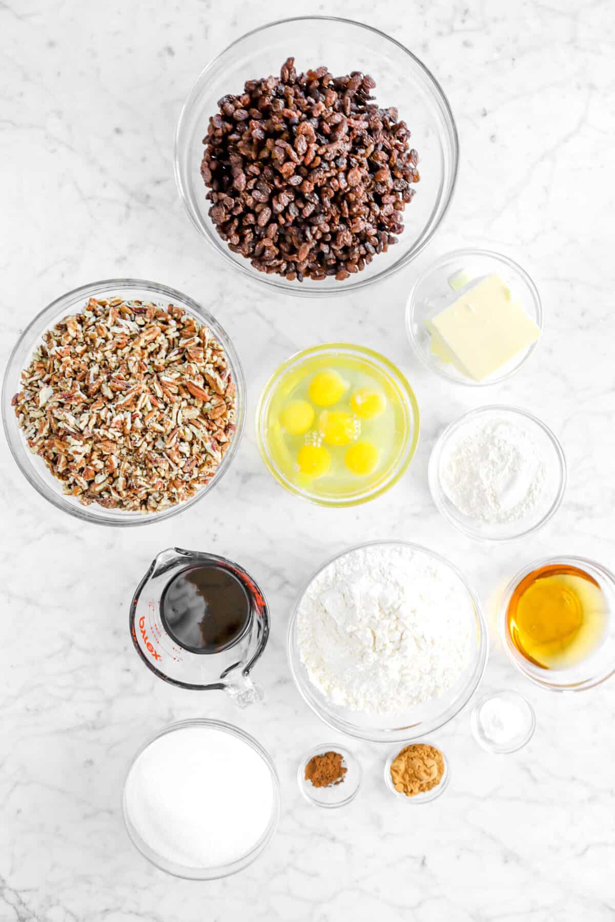 raisins, chopped pecans, butter, eggs, flour, molasses, bourbon, baking soda, cinnamon, cloves, and sugar in glass bowls on marble counter