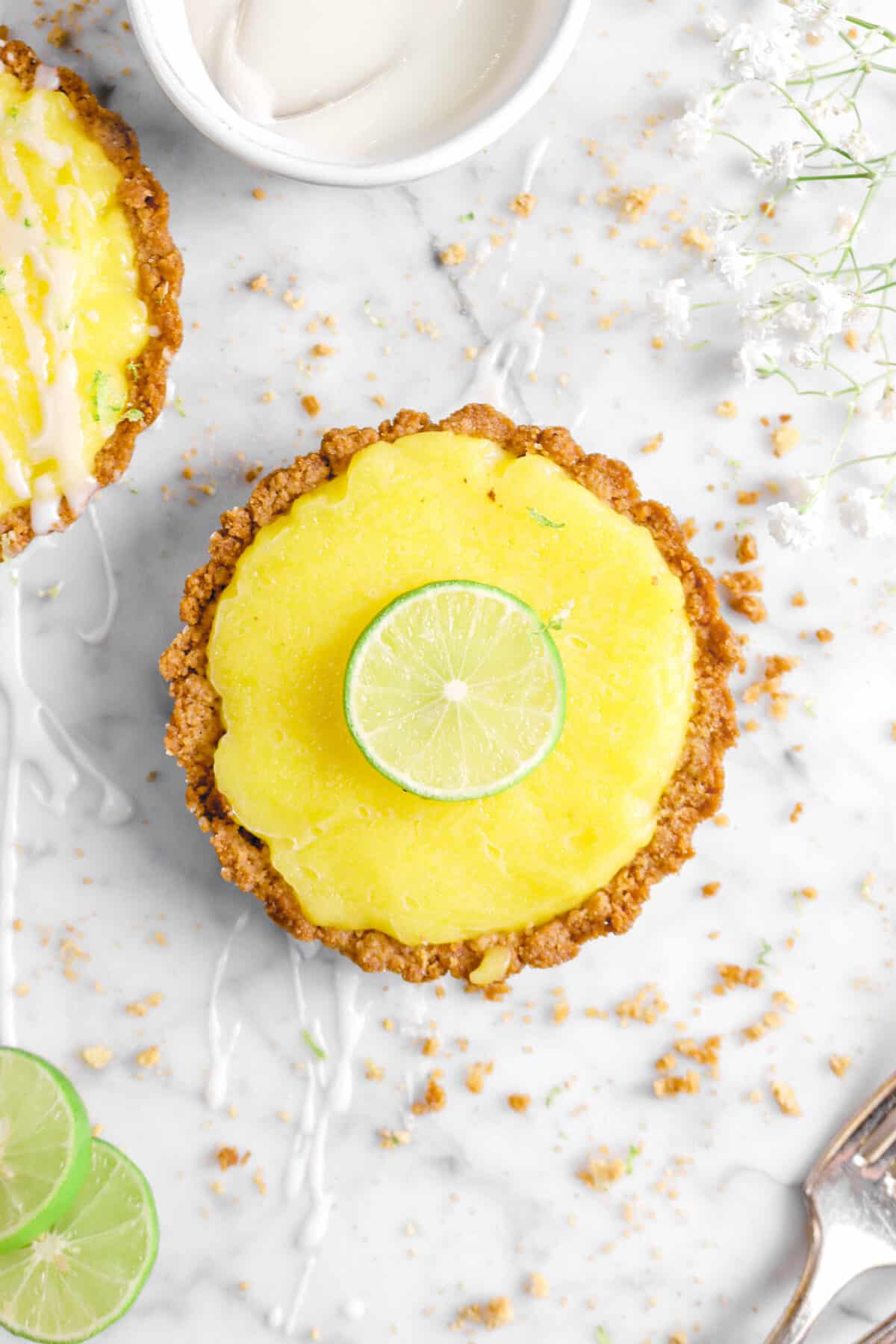 key lime pie with slice of key lime on top with flowers, crumbs, glaze, and fork
