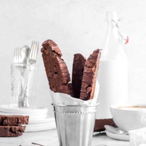 chocolate biscotti in metal cup with chocolate drizzle, a white napkin, mug of coffee, milk glass, and utensils behind