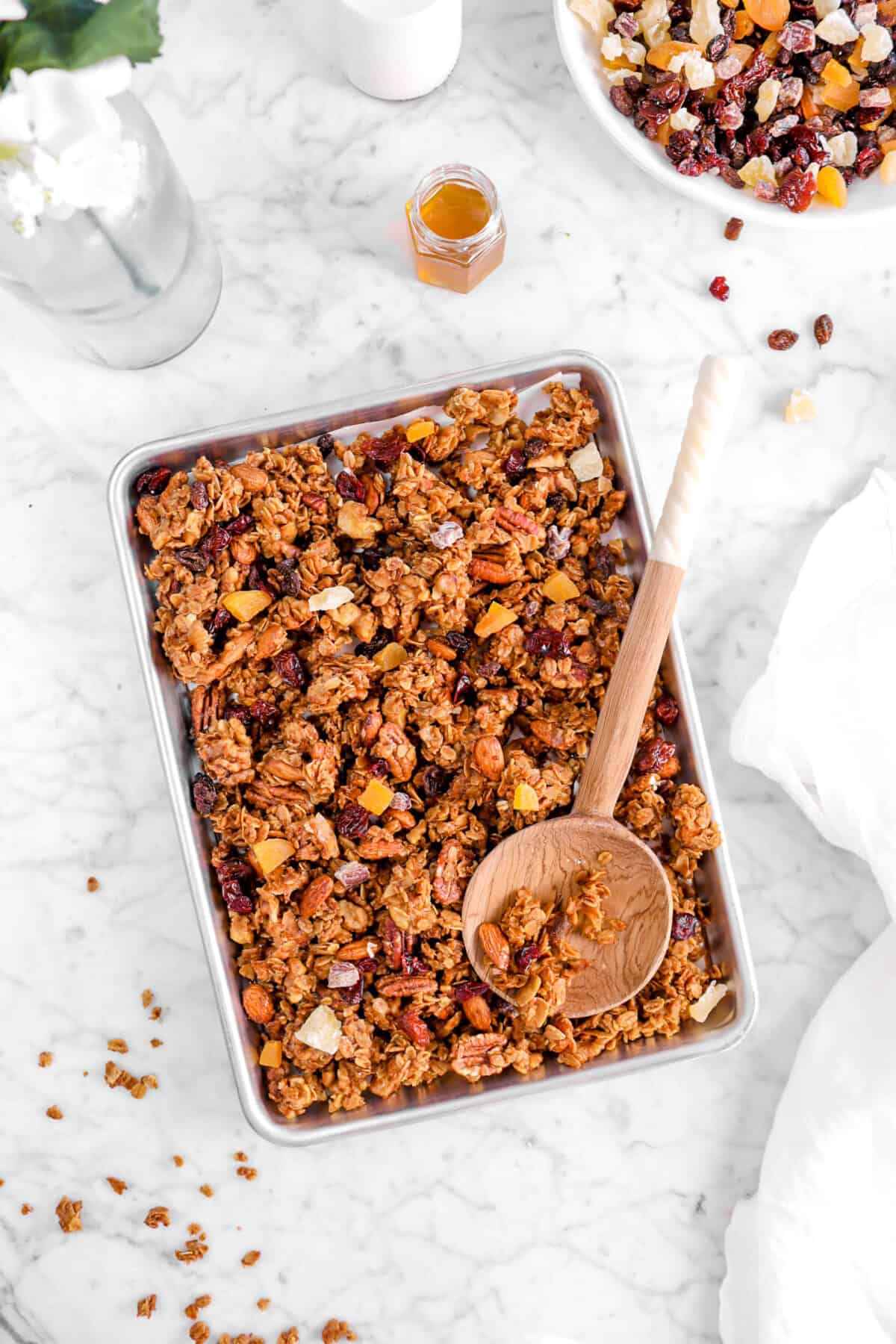 overhead shot of granola with wooden spoon, flowers, jar of honey, and bowl of fruit