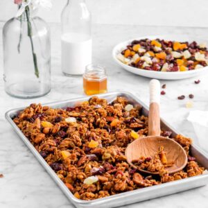 granola in sheet pan with wooden spoon, bowl of dried fruit, milk glass, honey pot, and flowers