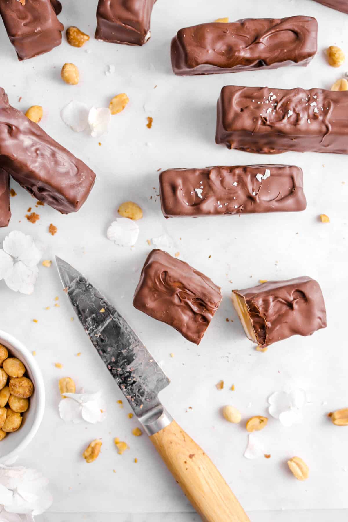 overhead shot of some snickers bars on parchment with one cut in half, a knife, peanuts, and flowers