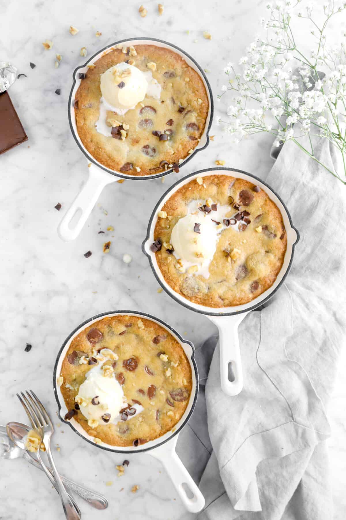 three white skillets of cookie with ice cream, chocolate curls, white flowers, and utensils