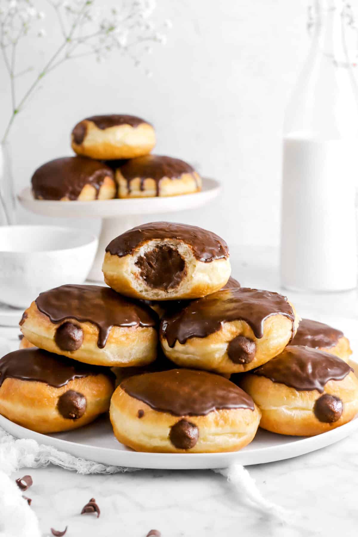 seven doughnuts on a white plate with chocolate glaze and a bite taken out of one with a bottle of milk and three doughnuts behind