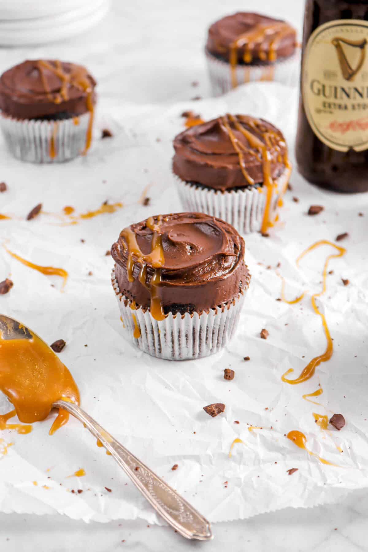 chocolate cupcake with caramel sauce drizzled on top with bottle of guinness behind, three cupcakes, and spoon of caramel