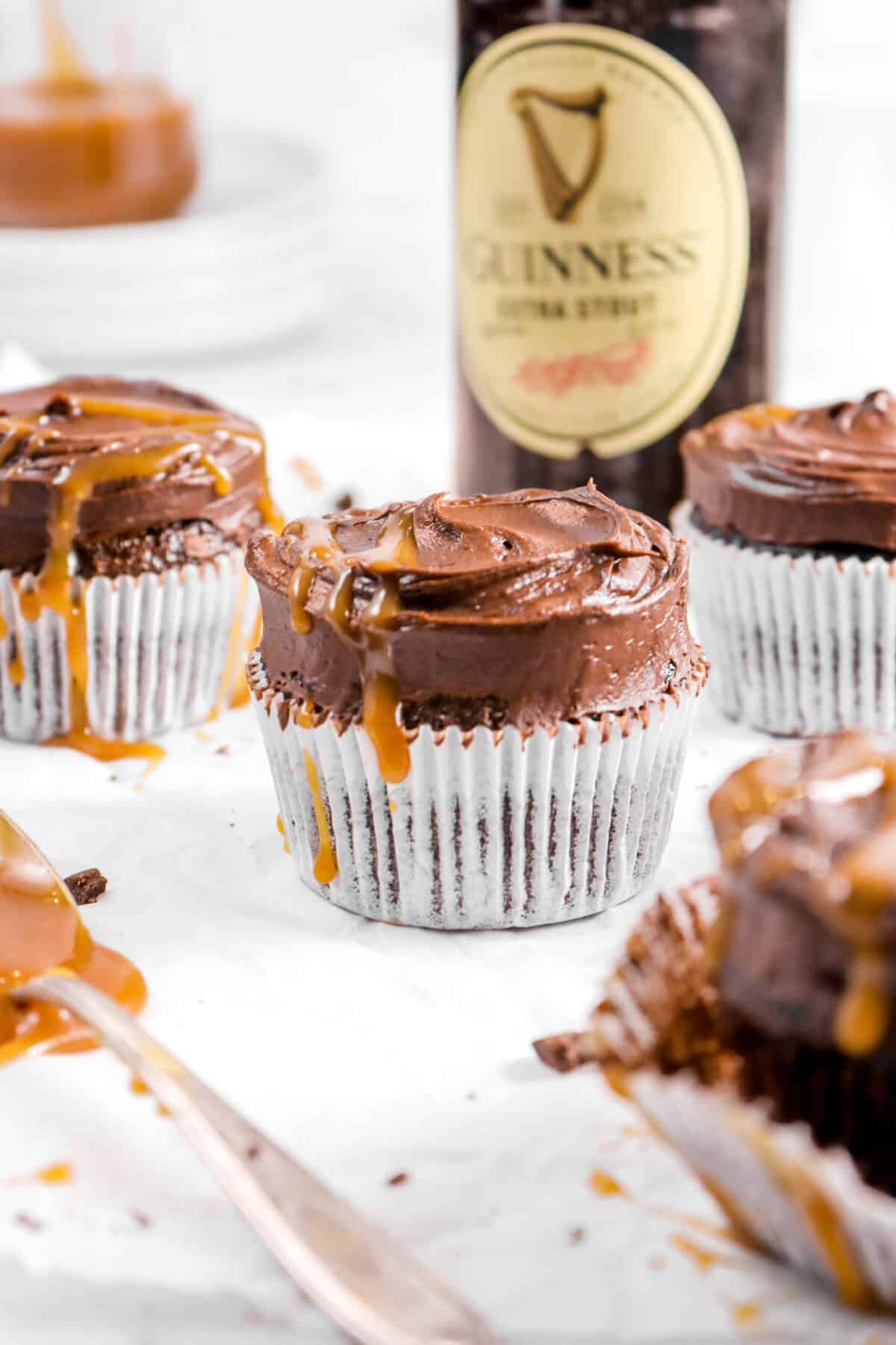 close up of guinness cupcakes with bottle of beer behind, spoon, and jar of caramel sauce on stacked plates