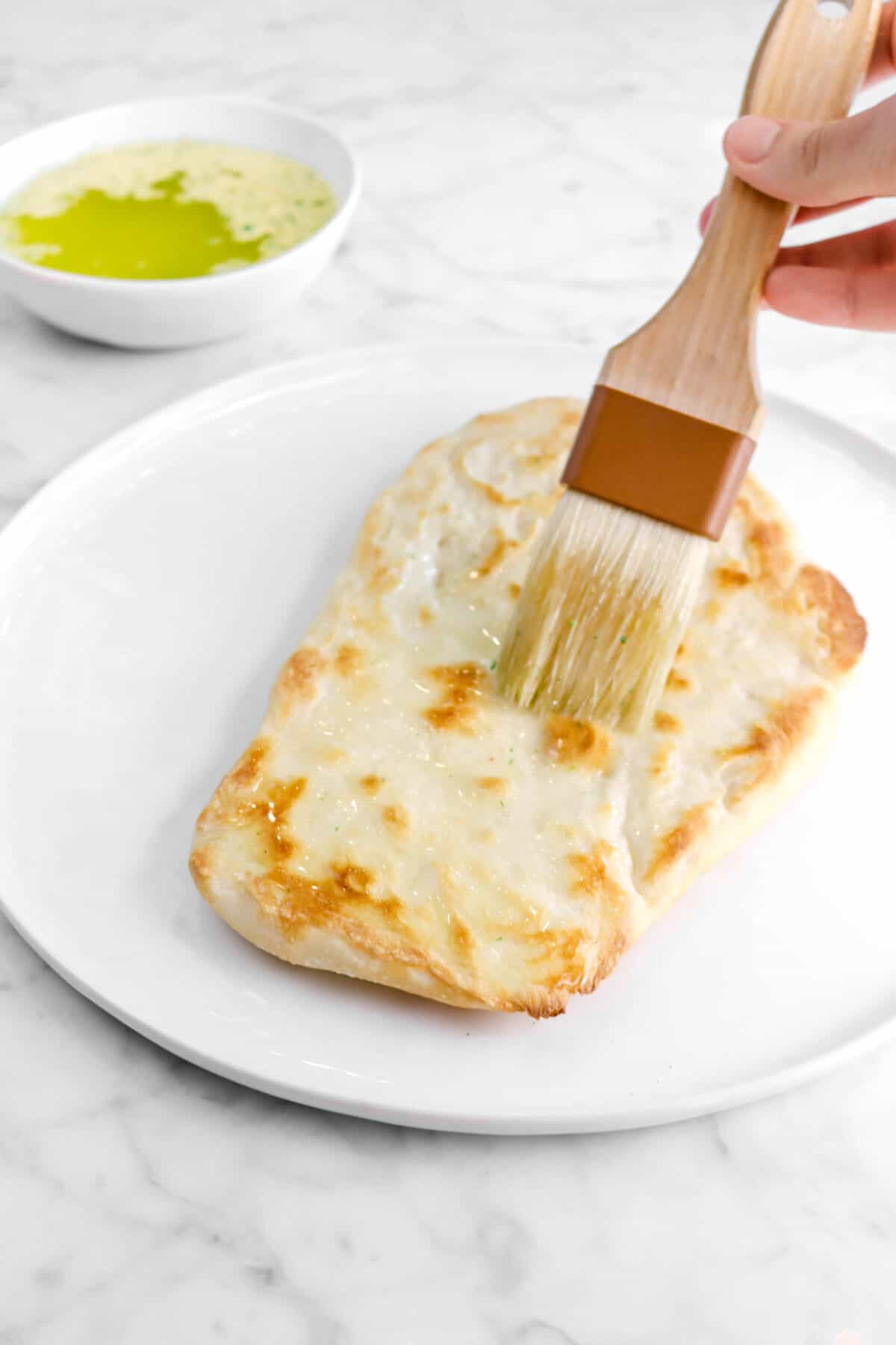 butter being brushed onto naan on a white plate with bowl of butter behind