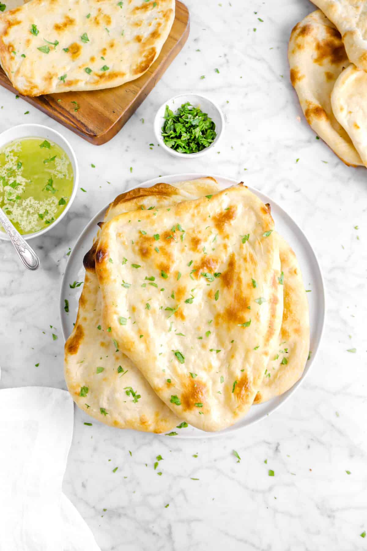 overhead shot of naan on white plate with chopped parsley on top, bowl of butter, bowl of chopped parsley, and more naan around