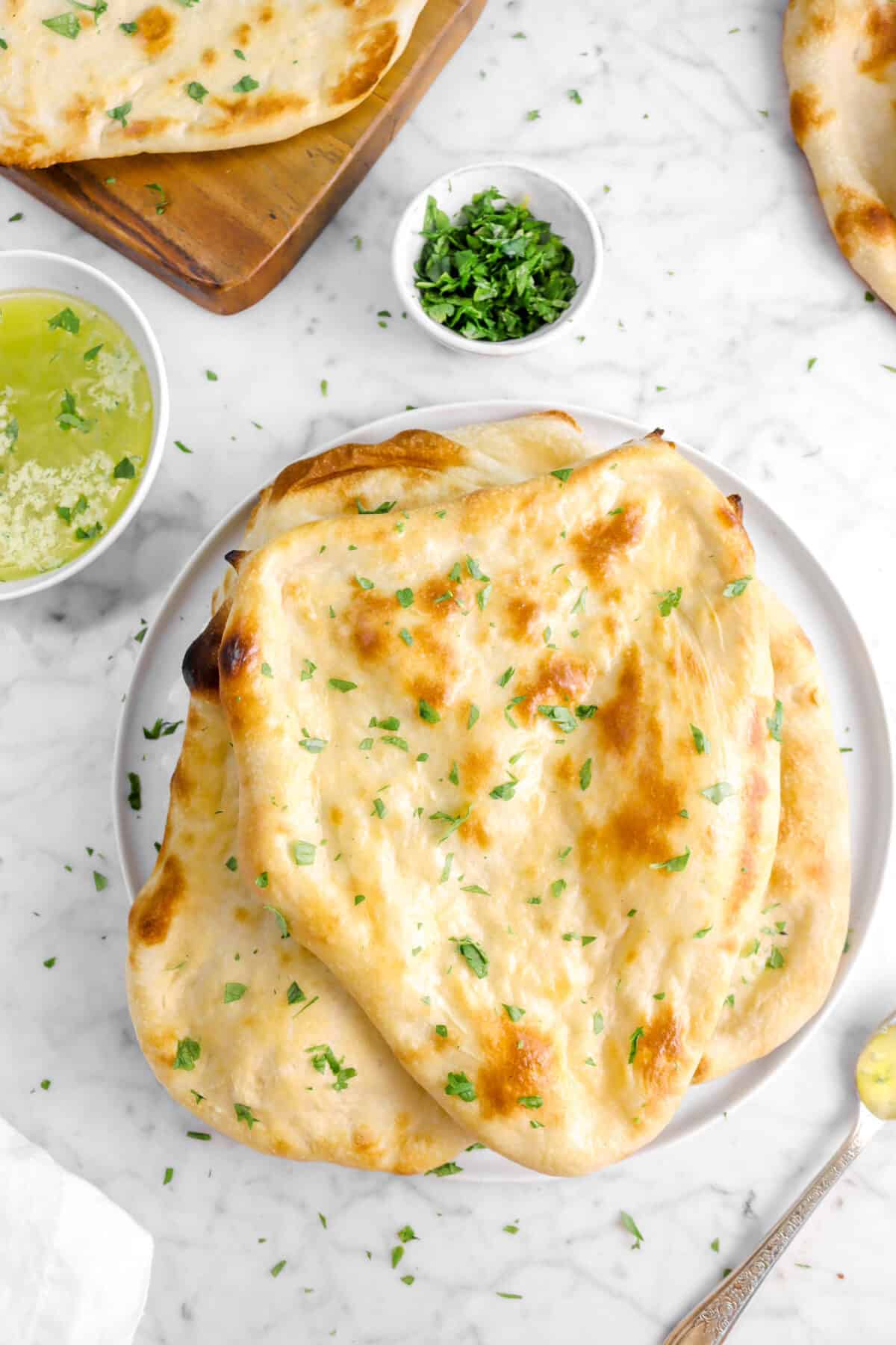 close up of naan stacked on a white plate with bowls of butter and chopped parsley