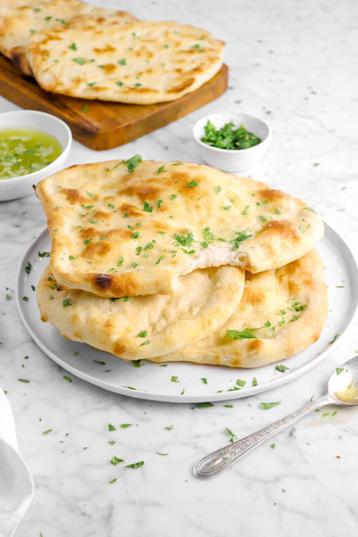 torn naan atop two others on a white plate with spoon, chopped parsley, and more naan behind