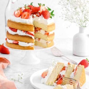 slice of strawberry shortcake layer cake being cut into with fork, full cake behind with jar of flowers