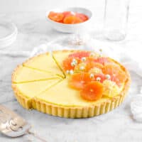 citrus tart with three slices laying next to it, a cake knife, white napkin, glass, and bowl of citrus slices