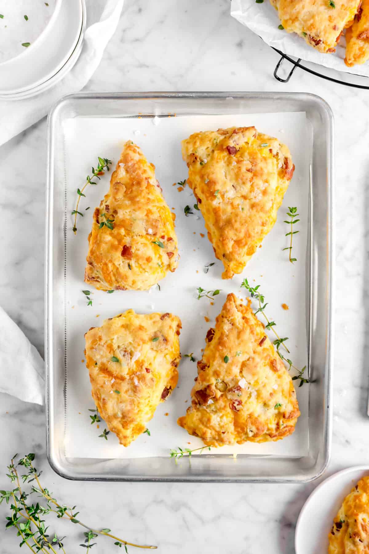 four scones on sheet pan with thyme sprigs, a napkin, plates, and more scones in the top right and bottom left corners of the image
