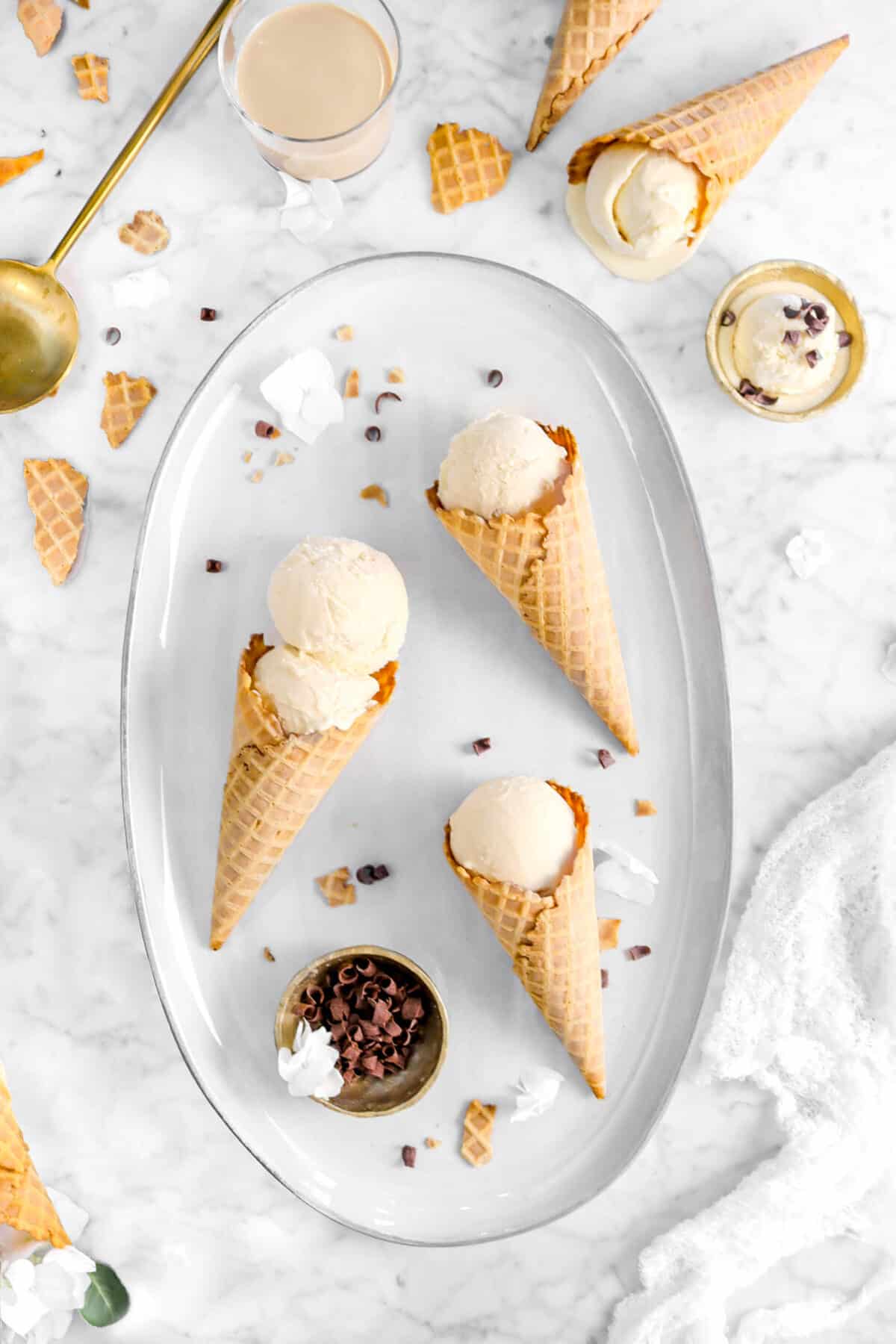 three cones of irish cream ice cream on oval serving board with a bowl of chocolate curls on it, ice cream cones around, a glass of irish cream, gold spoon, and bowl of ice cream